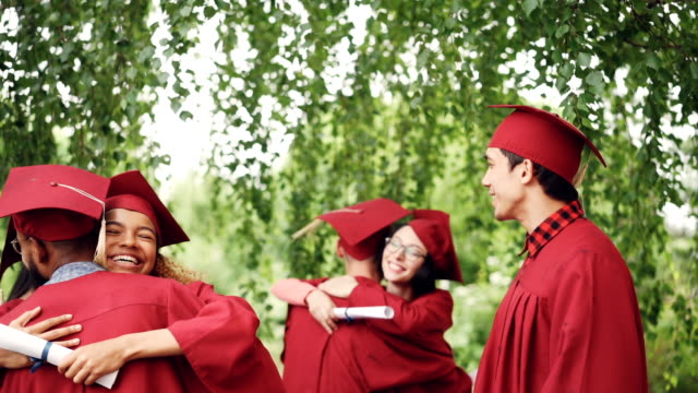 Happy-graduating-students-multi-ethnic-group-is-hugging-and-doing-high-five-after-graduation-ceremony,-girls-and-guys-in-mortar-boards-and-gowns-are-laughing-and-having-fun.
