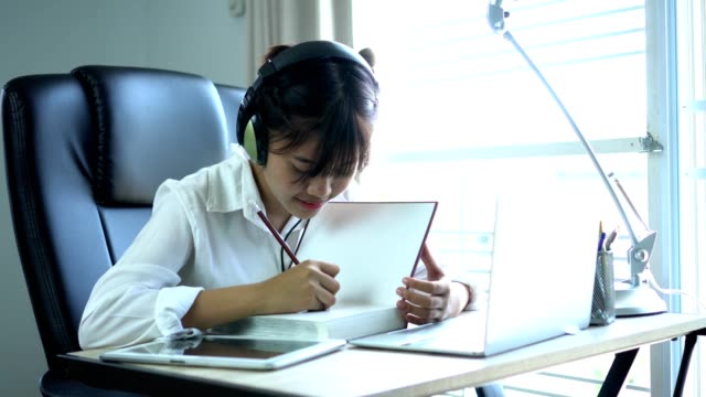 Student-Online-Studie-Lernkonzept:-schöne-Asiatin-hören-mit-Kopfhörern-und-Laptop,-sitzen-Lächeln-Schreiben-von-Notizen-im-Lehrbuch-an-ihrem-Schreibtisch-in-Haus-für-e-Learning-in-der-Bildungstechnologie