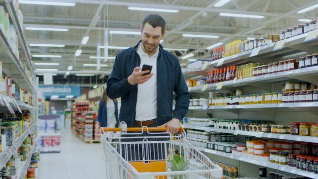 Im-Supermarkt:-gut-aussehend-Mann-nutzt-Smartphone,-lächelt-beim-stehen-im-Abschnitt-aus-der-Dose-waren.-Warenkorb-mit-gesunden-Lebensmitteln-im-Inneren-hat.-Andere-Kunden,-die-im-Hintergrund-laufen.
