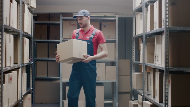 Trabajador-almacén-guapo-entra-en-almacén-con-una-caja-de-cartón-y-pone-en-un-estante.