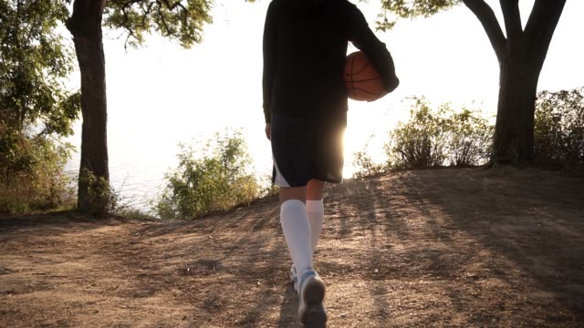 A-basketball-player-girl-comes-with-a-ball-in-her-hand,-coming-up-to-the-slope-with-trees-around.-Looks-at-the-sun-shining-over-the-sea.-Backside-view