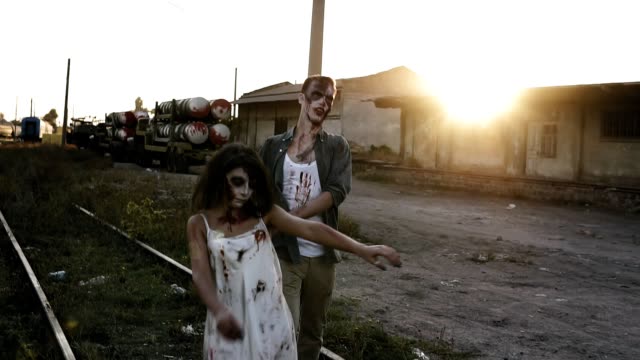 Halloween,-filming,-creepy-concept.-Creepy-zombie-man-and-woman-in-bloody-clothes-walking-by-railway-lines-outdoors-by-industrial-abandoned-place.-Sun-shines-on-the-background