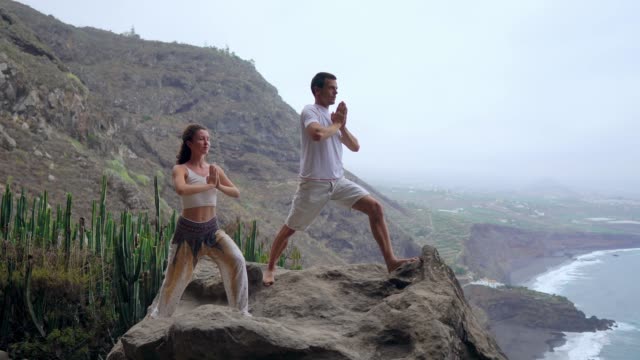 A-man-and-a-woman-standing-on-the-edge-of-a-cliff-overlooking-the-ocean-raise-their-hands-up-and-inhale-the-sea-air-during-yoga.