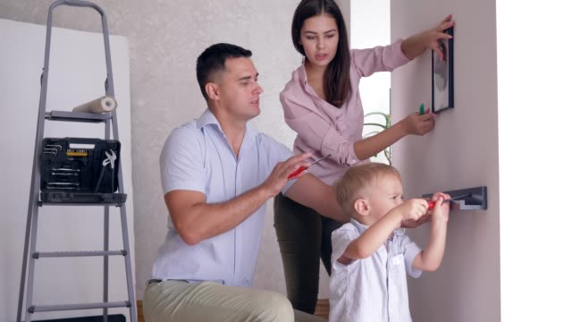 freundliche-Familie-Reparaturen-ausführen-und-kleinen-Sohn-hilft-Eltern-Regal-in-neue-Wohnung-an-Wand-befestigen