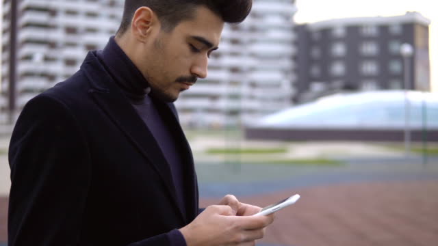 Young-urban-businessman-professional-in-black-suit-walking-in-street-using-app-texting-sms-message-on-smartphone