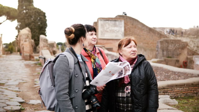 Joven-caucásica-mujer-excursión-guía-dar-detalle-en-las-históricas-ruinas-de-Ostia,-Italia-a-dos-turistas-senior-femenino