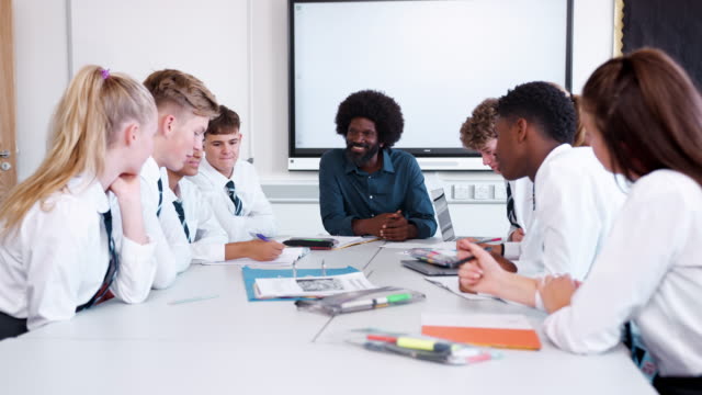 Male-High-School-Teacher-Sitting-At-Table-With-Teenage-Pupils-Wearing-Uniform-Teaching-Lesson