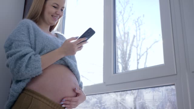 moderne-Schwangerschaft,-hübschen-schwangeren-Frau-mit-dicken-Bauch-sieht-in-smart-Handy-gegen-Fenster-im-Sonnenlicht-an-Wintertag