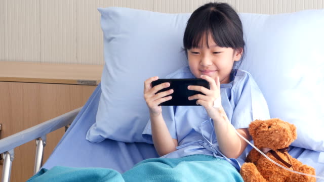 Kleine-Mädchen-liegt-auf-einem-Bett-im-Krankenhaus,-sie-Filme/Zeichentrickfilme/lustige-Videos-auf-Smartphone.-Menschen-mit-Technik,-Gesundheits--und-medizinische-Konzept.