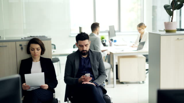 Candidatos-estresados-están-esperando-para-la-entrevista-de-trabajo-en-la-oficina-leyendo-el-cv-y-con-smartphone-mientras-manager-femenino-está-entrevistando-a-un-hombre-en-la-habitación.-Concepto-de-empresa-y-personal.