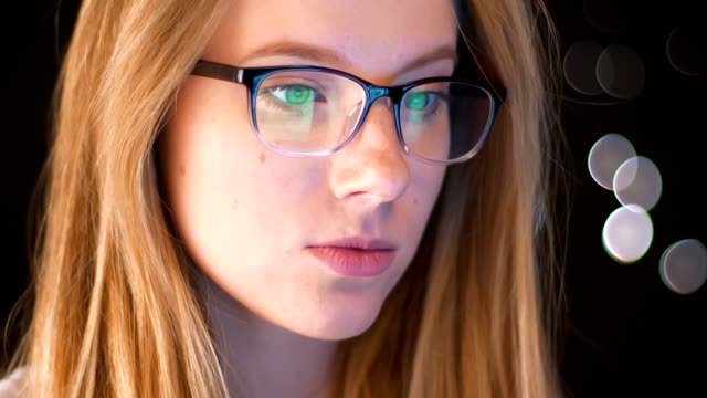 Reflection-of-green-screen-in-spectacles-on-caucasian-blonde-female,-who-is-standing-focused-on-using-device-and-being-relaed-on-dakr-background