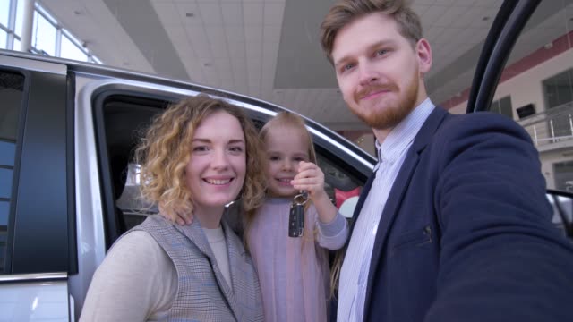 family-selfi-photo-in-auto-shop,-funny-couple-with-cute-kid-girl-with-keys-photographed-on-smartphone-near-new-purchased-automobile-in-car-sale-center