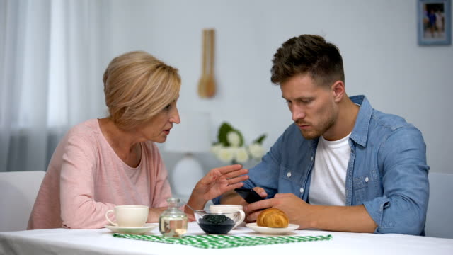 Worried-mother-talking-to-infantile-adult-son-playing-video-games-on-smartphone