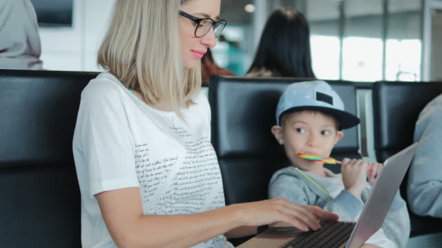 Mamá-joven-trabajando-en-la-computadora-portátil-en-el-aeropuerto-con-el-niño-disfrutando-de-piruleta.