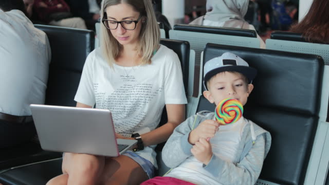 Young-mum-working-on-laptop-on-airport-with-child-enjoying-lollipop.