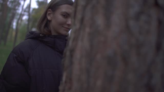 Pretty-girl-in-warm-coat-looking-out-from-behind-a-tree-smiling.-The-camera-moves-to-another-side-of-the-trunk
