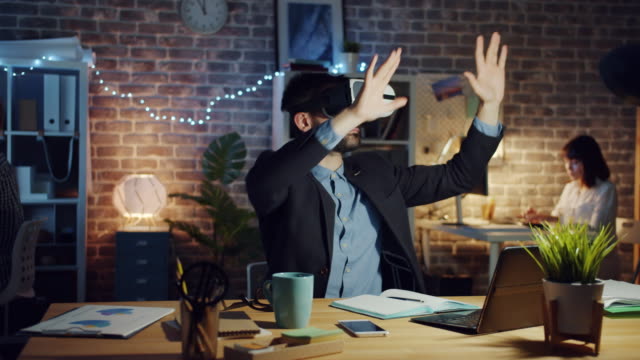 Mann-mit-Virtual-Reality-Brille-bewegt-Arme-sitzen-am-Schreibtisch-im-dunklen-Büro