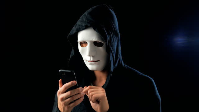Anonymous-enmascarado-estafador-en-la-capucha-exige-un-rescate-para-el-chantaje-utilizando-un-teléfono-inteligente.-Masked-Criminal-intimida-a-la-víctima-con-la-ayuda-de-amenazas-a-través-de-SMS-utilizando-el-teléfono-móvil.