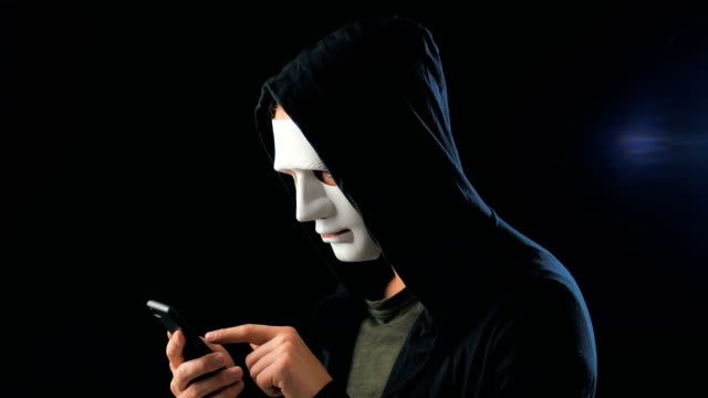 Anonymer-maskierter-Betrüger-in-Kapuze-fordert-ein-Lösegeld-für-Erpressung-mit-einem-Smartphone.-Maskierter-Krimineller-schüchtert-das-Opfer-mit-Hilfe-von-Drohungen-per-SMS-per-Handy-ein.