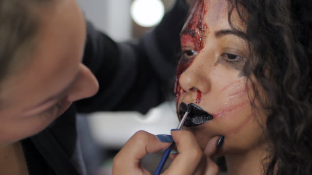 Make-up-artist-make-the-girl-halloween-make-upin-studio.Halloween-face-art.-Woman-applies-on-black-lipstick-with-brush-on-lips-of-latin-girl.-War-paint-with-blood,-scars-and-wounds.Slow-motion