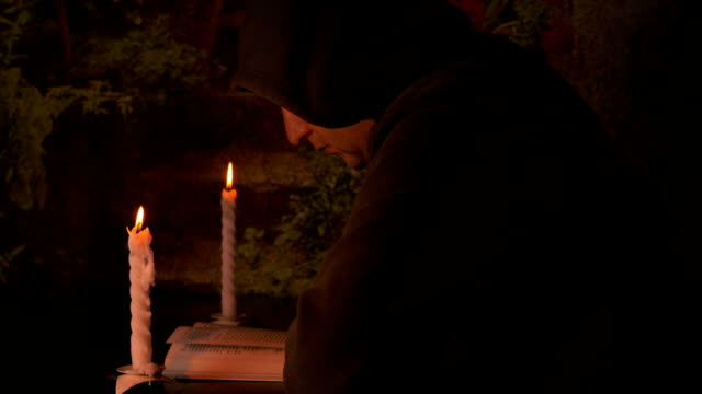 Burning-candle-in-a-small-white-candlestick-.-A-monk-in-a-hood-reading-a-book-.-Selective-focus.-Got-find-evrika-gestures.