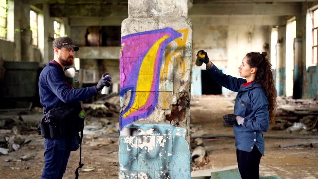 Slow-motion-of-two-serious-graffiti-artists-decorating-empty-industrial-building-with-beautiful-images-using-bright-aerosol-paint.-Creative-teamwork,-people-and-art-concept.
