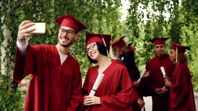 Cheerful-couple-of-graduating-students-is-taking-selfie-using-smartphone,-young-man-and-woman-are-holding-diplomas,-looking-at-smart-phone-camera-and-smiling.