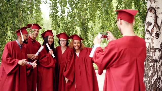 Graduating-student-is-recording-video-of-his-friends-in-gowns-holding-diplomas,-waving-hands-and-posing-looking-at-smartphone-camera.-People-and-technology-concept.
