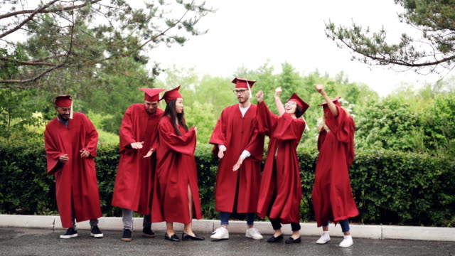 Slow-motion-of-multi-ethnic-group-of-students-in-gowns-and-mortar-boards-dancing-and-laughing-outdoors-on-campus-on-graduation-day-celebrating-event.
