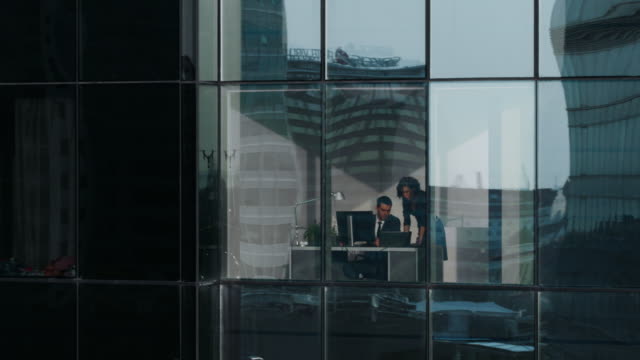 Imágenes-aéreas-de-fuera-de-los-rascacielos:-empresario-y-empresaria-hablando-de-negocios-sentado-en-el-escritorio-en-la-ventana-de-la-oficina.-Toma-vuelo-del-distrito-financiero-y-empresarios-trabajando-en-la-gran-ciudad.