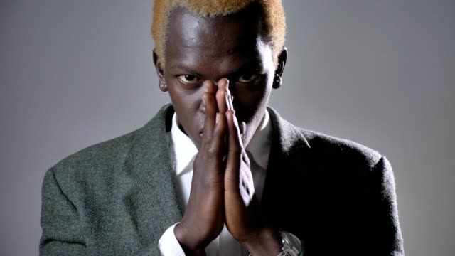 Junge-afro-amerikanische-blonden-Mann-im-Anzug-Händchenhalten-und-beten,-suchen-in-der-Kamera-auf-grauem-Hintergrund-isoliert