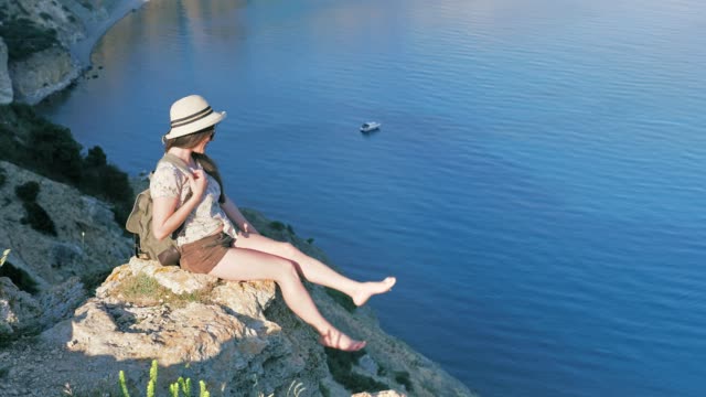 Bescheidenen-jungen-weiblichen-Reisenden-mit-Rucksack-auf-Felsen-sitzen-und-spielen-ihre-nackten-Füße-bei-Sonnenuntergang
