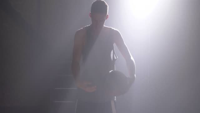 Jugador-de-baloncesto-realizando-diferentes-trucos-con-la-bola,-girando-y-agitando,-de-pie-en-el-gimnasio-oscuro-con-niebla