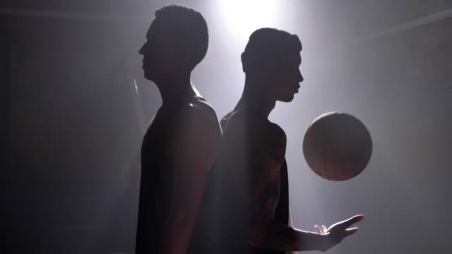 Zwei-Basketball-Spieler-Silhouette-stehen-Rücken-an-Rücken-in-Raum-mit-Rauch-und-Flutlicht