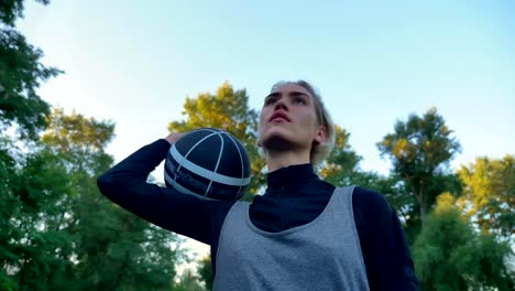 Selbstbewusste-junge-Frau-Holding-Basketball-und-wir-freuen-uns,-stehen-im-Park-mit-blauen-Himmel-oben,-niedrigen-Winkel