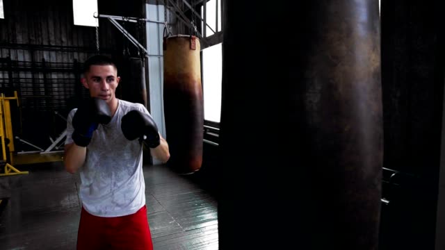 Boxer-hombre-caucásico-en-camiseta-y-camisas-rojas-ejercitándose-en-el-gimnasio.-Practicando-patadas-y-puches-con-saco-de-boxeo.-Boxeo-en-el-interior-del-estudio