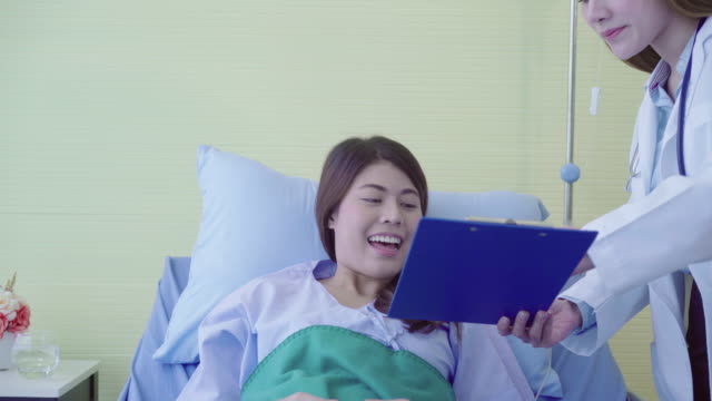 Beautiful-smart-Asian-doctor-and-patient-discussing-and-explaining-something-with-clipboard-in-doctor-hands-while-staying-on-Patient's-bed-at-hospital.-Medicine-and-health-care-concept.