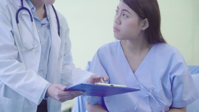 Schöne-intelligente-asiatischen-Arzt-und-Patient-diskutieren-und-etwas-mit-Zwischenablage-in-Händen-der-Arzt-während-des-Aufenthaltes-am-Bett-des-Patienten-im-Krankenhaus-zu-erklären.-Medizin-und-Gesundheitswesen-Konzept.