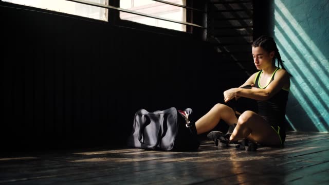Panning-young-beautiful-woman-sitting-on-floor-and-wrapping-hands-with-black-boxing-wraps-in-club.