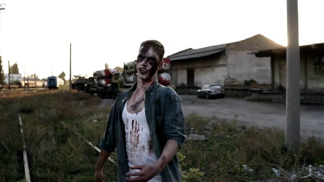 Creepy-zombie-man-in-bloody-clothes-walking-by-railway-lines-outdoors-with-an-industrial-abandoned-place-on-the-background.-Halloween,-filming,-creepy-concept