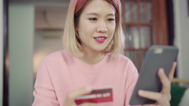 Beautiful-Asian-woman-using-smartphone-buying-online-shopping-by-credit-card-while-wear-sweater-sitting-on-desk-in-living-room-at-home.-Lifestyle-woman-at-home-concept.