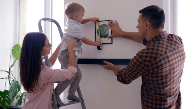 hübsches-Kind-mit-Maßband-hilft-Mutter-und-Vater-hängen-Regal-mit-Bild-an-die-Wand-nach-der-Reparatur-in-Wohnung