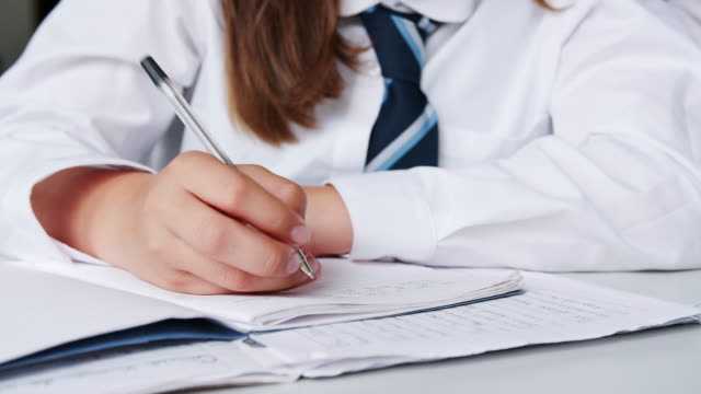 Close-Up-Of-Female-High-School-Students-Wearing-Uniform-Writing-In-Exercise-Book-At-Desk