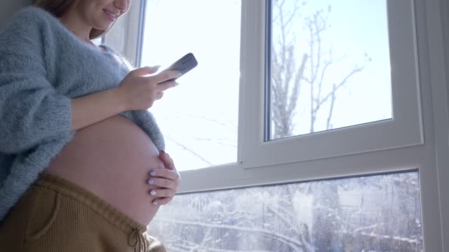 moderne-zukünftige-Mutter,-schöne-Schwangerschaft-Frau-mit-dicken-Bauch-mit-Mobile-Smartphone-zu-Hause-gegen-Fenster-im-Sonnenlicht-an-Wintertag