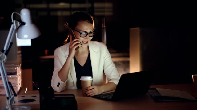 Attractive-female-entrepreneur-is-talking-on-mobile-phone-and-using-laptop-working-in-office-late-at-night.-Modern-technology,-hard-working-youth-and-communication-concept.