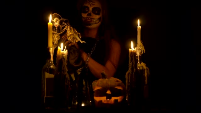 Bruja-de-Halloween-con-maquillaje-de-cráneo-hace-vudú-sostiene-cuchillo-y-wispering-hechizo-mágico-cadenas-de-calabaza-y-velas