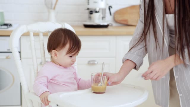 Young-mother-feeding-her-baby-daughter-fruit-puree-and-wiping-her-face-with-tissue-in-domestic-kitchen.-Baby-girl-learning-to-eat-with-spoon-sitting-in-highchair