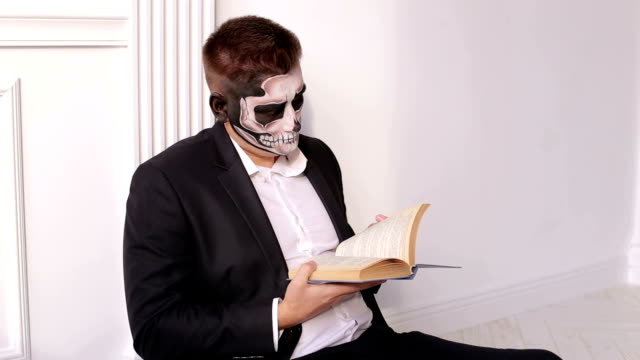 Un-hombre-con-un-maquillaje-terrible-en-forma-de-cráneo-está-sosteniendo-un-libro.-Víspera-de-Todos-los-Santos