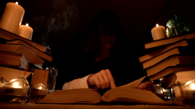 Mittlere-Nahaufnahme-Mädchen-Magier-in-einer-Kapuze-in-einem-dunklen-Raum-bei-Kerzenschein-und-auf-der-Suche-nach-einem-Zauber,-der-ein-Buch-umdreht.-Niedriger-Schlüssel.