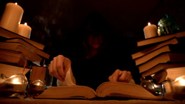 Medium-close-up-girl-magician-in-a-hood-in-a-dark-room-by-candlelight-and-looking-for-a-spell-turning-over-a-book.-Low-key.